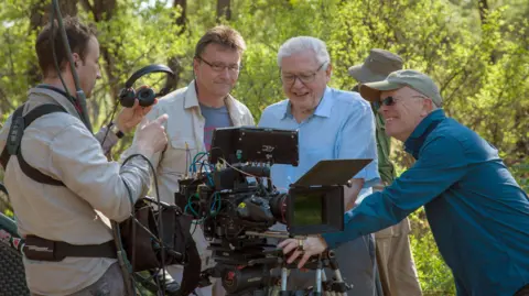 BBC Studios Sir David Attenborough and others looking at a film camera in a green woodland with the sun shining