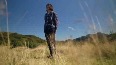 Botanist Heather Garrett stood in a field with long yellow grass. She's wearing dark trousers, a blue patterned long sleeved top and has a short dark bob and glasses