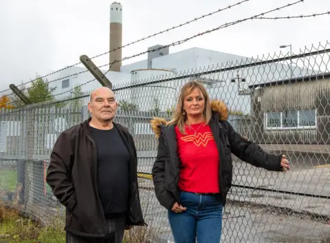 BBC / Jon Parker Lee Runcorn residents George Parker, garage owner and Mandy Royle stand solemnly infront of a fence. In the background is the Runcorn waste incinerator, which backs onto their properties.   