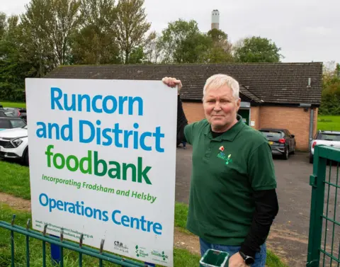 BBC / Jon Parker Lee  Eddie Thompson, manager of the Runcorn Foodbank at the charities main distribution centre, standing next to sign. Incinerator can be seen in the distance. 