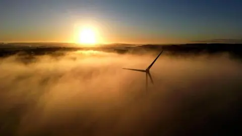 Reuters Wind turbine seen through fog as the Sun rises in background