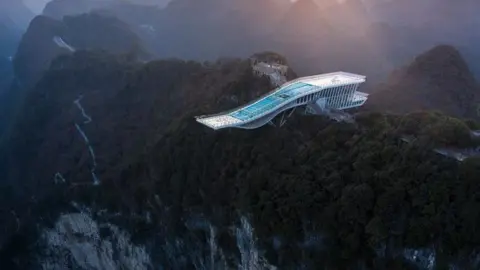 Bowen Hou @AUPH A futuristic platform reaches out from a forested cliff edge over a massive sinkhole. Mountains lit by a low sun stretch out in the background