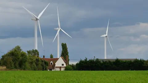 John Keeble/Getty Images Wind farm turbines are seen behind a residential house in Bradwell-on-Sea, United Kingdom, July 2024