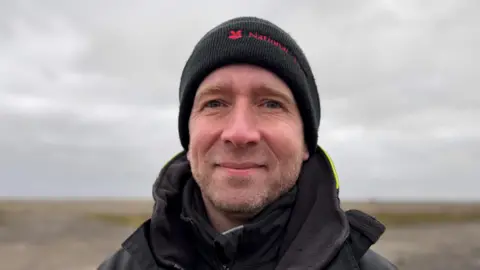 Jo Black/BBC Matt Wilson smiles at the camera while standing on a shingle beach. He is wearing a black coloured beanie hat and a black coat. He has light coloured stubble.