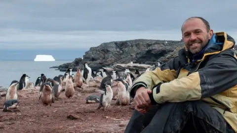 BAS Derren Fox is sitting on the ground smiling at the camera. He has a beard and moustache and is wearing a black and yellow anorak, jeans and walking boots. He is surrounded by dozens of penguins and there is a cliff and the sea behind him. Large icebergs are in the sea