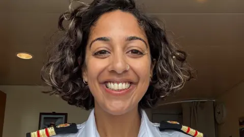 BAS Dr Nisha Mistry is smiling at the camera. She has dark, wavy hair just above shoulder length. She is wearing earrings and has a ring through her nose. She is wearing a white shirt with epaulettes