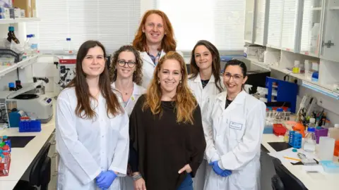 Weizmann Institute of Science Group of six scientists - five women and a man - taking a group picture, most wearing white lab coats in a laboratory setting.