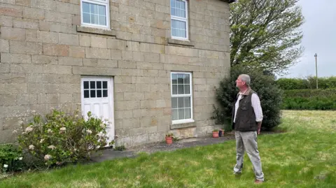 BBC Martin Beal is looking at his house which is a Georgian stone built farmhouse with a white door and windows. He is a man, about 60 years of age, wearing a waist coat, white shirt and light coloured trousers.