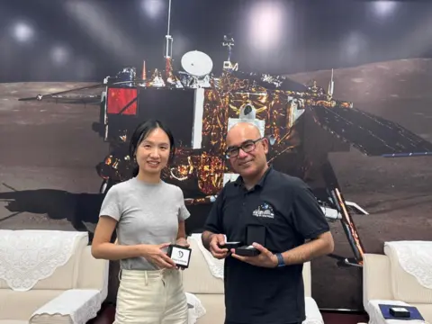 Mahesh Anand Mahesh Anand and a woman working with the Chinese space agency standing in front of a large picture of a spacecraft with black arms and gold material on the outside. They are standing in front of white sofas and holding small boxes. The boxes have white labels with the Chinese space agency logo. 