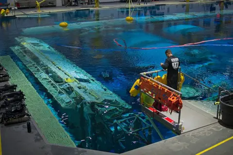 Kevin Church/BBC Wide of support team member standing on a platform overlooking the water of the pool. Some of the modules of the space station are clearly visible below the surface. There is a row of scuba equipment to the left of the picture and there are red and blue