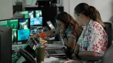 Kevin Church/BBC Trainers in the control room are leaning in to a microphone and communicating with the astronauts. There is a bank of screens in front of them. The woman on the left is wearing a blue flowery shirt and has brown hair and glasses. The woman on the right is wearing a white shirt with red stylised flowers. The dive is taking place on