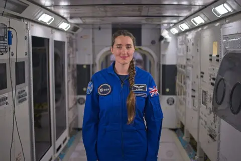 Kevin Church/BBC Picture of astronaut Rosemary Coogan in the replica of the International Space station at Johnson Space Centre. She is in the middle of the shot and has brown hair with a long plait that hangs down over her left shoulder. She is wearing a blue jumpsuit with a Union Jack on her left arm her name, and the European Space Agency logo on the front and the badge for her astronaut group