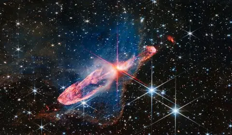 NASA A small bright red star in the centre in the process of forming as it illuminates a thick swirling  ring of gas around it a deep red. In the background are a field of stars