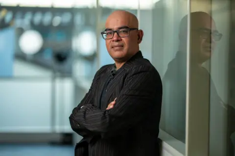 Tony Jolliffe/BBC News Professor Mahesh Anand stands with his arms folded and his back to a reflective screen in a corridor. 
