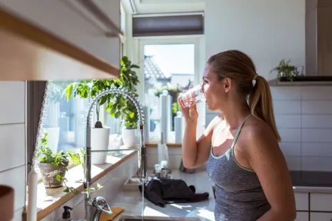 Getty Images A woman in a vest top with blonde hair tied at the back drinks a glass of water standing by a kitchen sink. The white-tiled kitchen has pots of green plants on the window sill and has a big glass window allowing sunlight into the kitchen. 