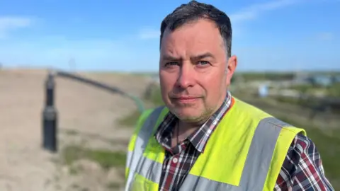 David Balmer, with short dark hair and stubble, wears a checked shift and high visibility vest.  He's standing in front of a methane extraction pipe on an active landfill site.