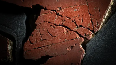 Tony Jolliffe/BBC News A close up of piece of red plaster, made up of several smaller pieces, with the letters F E C I T in capitals clearly visible with a line drawn in a square around three sides of the word. The left hand side of the plaster, where the name of the person or group who made the piece of art should be, is missing. 