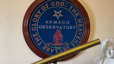 A large Armagh Observatory crest with a wooden frame on a wall. A shiny brass telescope on a shelf under the crest, along with a with bust of a Georgian era man.