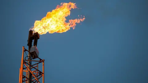 Karol Serewis/Getty Images An orange tower on the left of the picture, a orange flame is being emitted with black smoke trailing left to right. The tower stands against a blue sky. The flame is produced by flaring from oil production