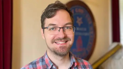 A close-up of Dr Rok Nežič smiling at the camera in Armagh Observatory. He has short, brown hair and a beard. He is wearing glasses and a red and blue checked shirt.
