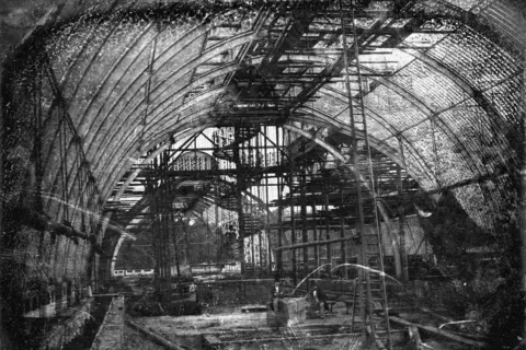 RBG Kew A black and white image taken inside the Palm House, a glass and iron building in Kew Gardens. It shows it part way though construction. Scaffolding and tall ladders run from the floor to the ceiling.