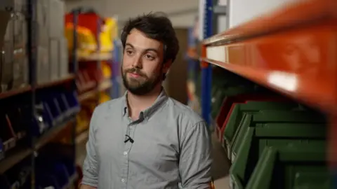 BBC News A man wearing a grey shirt stands in a corridor, with orange and green files around him.  