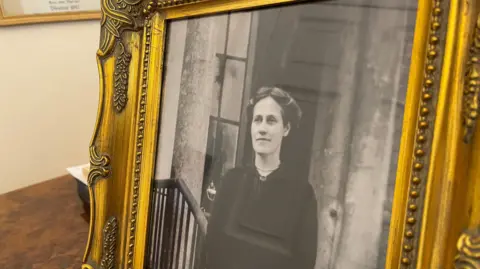 A framed black and white photo of Theresa Hardcastle. She has long hair which has been parted and tied up around her head. She is wearing a black dress and is standing on the steps of a large building, leaning one arm on a bannister.