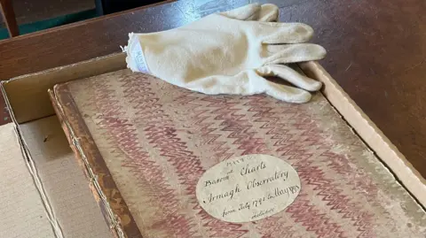 BBC A close-up of a very old book containing records of weather observations from Armagh Observatory from July 1795 to May 1798. The book, which has a hand-written cover and is decorated with wavy red lines, is in a cardboard box on a wooden desk. A white glove used for handling the delicate paper is lying on the book.
