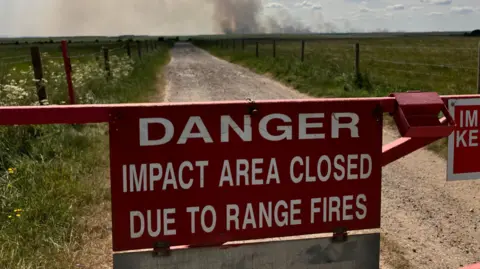 BBC A large red warning sign sits on a red barrier across a fenced dirt track that runs through a countryside plain. Large white writing warns