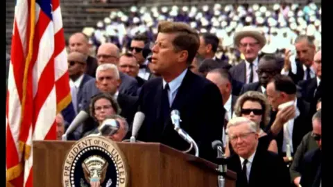 NASA JFK standing at a podium, with the seal of the US President upon which four microphones stand, an American flag to his right and massed crowds behind him. He appears young and energetic