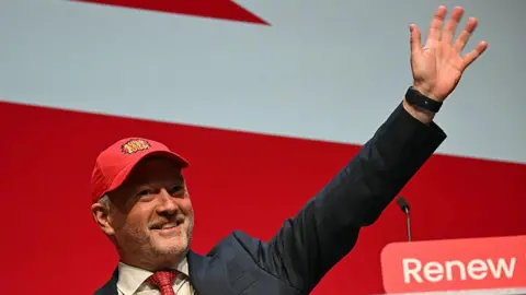 Getty Images A smiling Steve Reed in suit and tie waves, wearing a red baseball cap embroidered with the words 