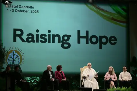 Getty Images On a stage in his summer residence, Castel Gandolfo, the Pope dressed in white, speaks to an audience. Four other speakers sit beside him, listening intently.