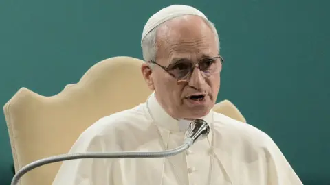 Getty Images Dressed in a white robe and white skullcap, Pope Leo speaks into a microphone while seated on a cream coloured chair, at the Raising Hope conference near Rome today