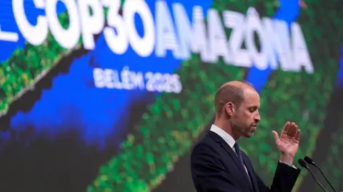 Aaron Chown/Getty Images Side view of Prince of Wales speaking in front of a lectern. He wears a dark blue suit and a purple tie. Behind him the words 