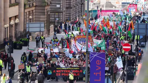 PA Media A wide overhead view of a large climate march filling a city street in Glasgow. Hundreds of people carry banners, placards and flags, including a large front banner reading “A Better World Is Possible.” Many signs reference climate justice, Gaza, and environmental protection. Police officers line the route, and tall buildings rise on both sides of the street as the crowd stretches far into the distance.