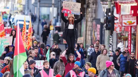 PA Media A large crowd of protesters marches down a city street. At the centre, a woman wearing a hat stands on someone’s shoulders and holds a sign above her head that reads, “There’s no wealth on a dead planet.” People around her carry flags and placards with various climate-justice messages. The street is busy with demonstrators, some in bright pink steward vests, and shops and buildings line the background.