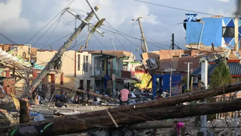Ricardo Makyn/AFP/Getty Images The aftermath of Hurricane Melissa is seen. Power lines, trees and houses have fallen down across a street. Rubble is strewn everywhere. The houses that remain are a mixed of brown, yellow and bright blue. A man in a pink top attempts to cycle through the debris.